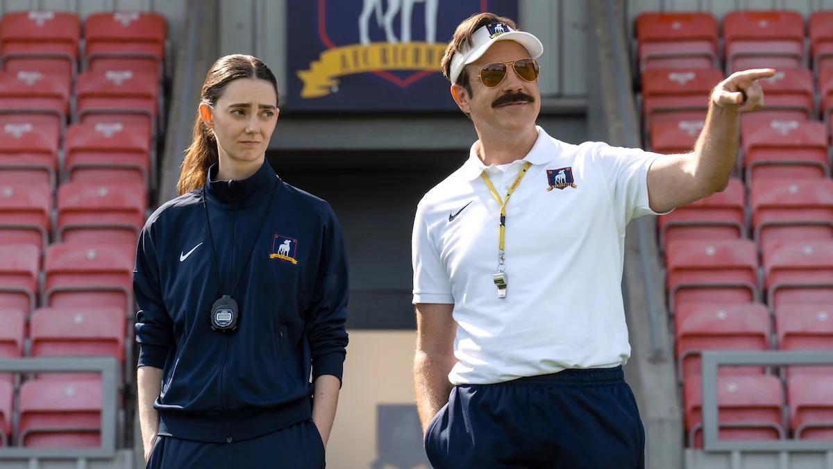Ted Lasso pointing out at the pitch as Tanya Reynolds looks on skeptically